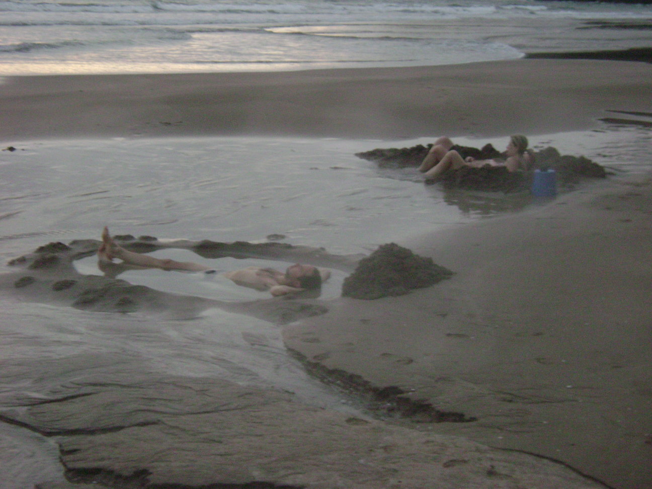 Downward view of a person laying in a watery hole on a beach, with a couple people laying in a sandy pit near them
